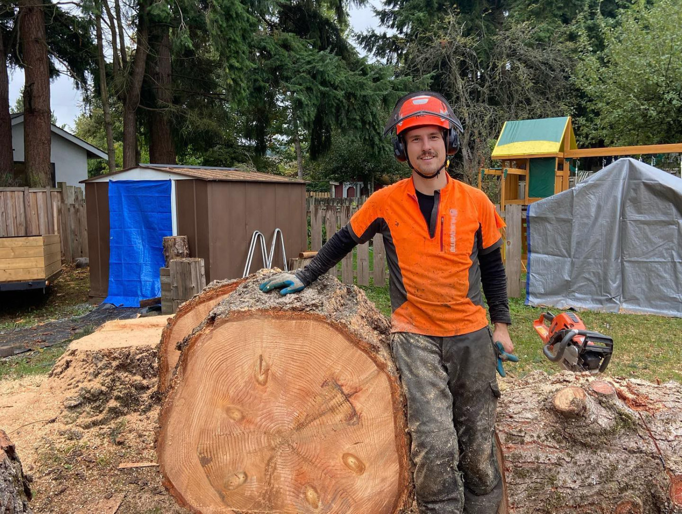 Matt Hardman, Owner and Operator of Summit Tree Care, standing next to a freshly cut large tree round. Matt is wearing full high-vis safety gear and smiling.